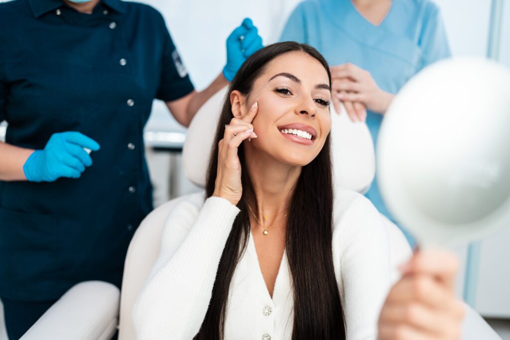 Beautiful and happy young woman sitting in a medical chair, smiling as she looks in the mirror, satisfied after a successful beauty treatment such as microneedling, dermaplaning, or hyaluronic acid fillers.