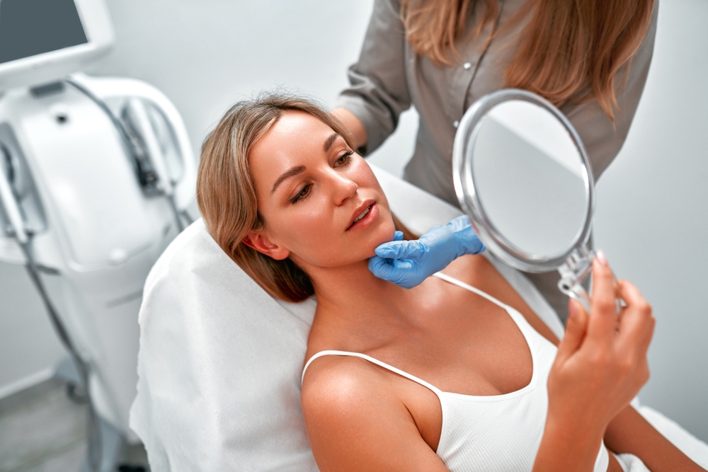 Cosmetologist consulting a female client in a beauty clinic as she looks in the mirror, preparing for a skincare procedure such as SmartXide DOT laser resurfacing for skin rejuvenation. Cosmetologist consulting a female client in a beauty clinic as she looks in the mirror, preparing for a skincare procedure such as SmartXide DOT laser resurfacing for skin rejuvenation.